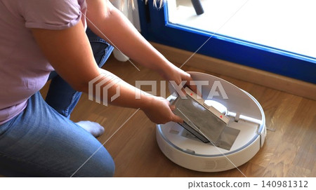 Woman removes dust bag from robotic vacuum cleaner while kneeling on wooden floor near a blue-framed window in a bright indoor setting Woman removes dust bag from robotic vacuum cleaner while kneeling on wooden floor near a blue-framed window in a bright indoor setting 140981312