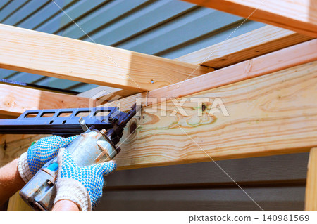 Contractor uses air pneumatic gun to nailing in wooden beams while working outdoors in residential area. Contractor uses air pneumatic gun to nailing in wooden beams while working outdoors in residential area. 140981569
