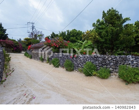 Vibrant bougainvillea blooms beyond the beautiful white sand paths and traditional stone walls of Taketomi Island. Vibrant bougainvillea blooms beyond the beautiful white sand paths and traditional stone walls of Taketomi Island. 140982843