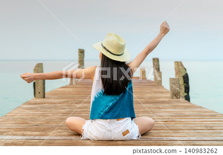 Rear view of a happy woman stretching her arms while sitting on a wooden pier overlooking calm sea. Freedom, travel, peaceful vacation, tropical lifestyle, wellness, relaxation. Summer holiday. Rear view of a happy woman stretching her arms while sitting on a wooden pier overlooking calm sea. Freedom, travel, peaceful vacation, tropical lifestyle, wellness, relaxation. Summer holiday. 140983262