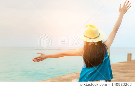 Rear view of a happy woman stretching her arms while sitting on a wooden pier overlooking calm sea. Freedom, travel, peaceful vacation, tropical lifestyle, wellness, relaxation. Summer holiday. Rear view of a happy woman stretching her arms while sitting on a wooden pier overlooking calm sea. Freedom, travel, peaceful vacation, tropical lifestyle, wellness, relaxation. Summer holiday. 140983263