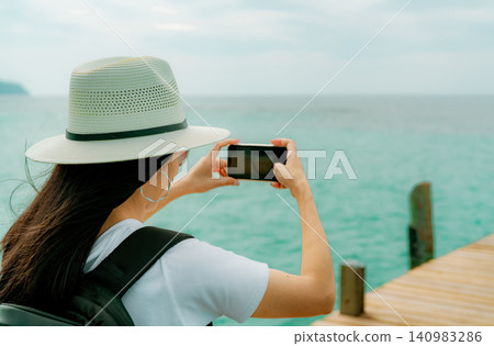 A female traveler using a smartphone to photograph the turquoise tropical sea from a wooden pier during a sunny summer vacation, capturing travel memories, mobile photography, and coastal tourism A female traveler using a smartphone to photograph the turquoise tropical sea from a wooden pier during a sunny summer vacation, capturing travel memories, mobile photography, and coastal tourism 140983286