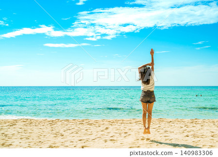 Young woman traveler stretching on a tropical sandy beach beside turquoise ocean beneath blue sky, expressing wellness, fitness, freedom, relaxation, summer vacation, and healthy travel lifestyle. Young woman traveler stretching on a tropical sandy beach beside turquoise ocean beneath blue sky, expressing wellness, fitness, freedom, relaxation, summer vacation, and healthy travel lifestyle. 140983306