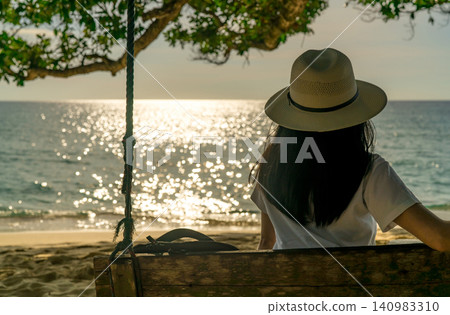 Back view of a woman relaxing on a wooden beach swing beneath tropical tree at sunset, enjoying the sparkling ocean, peaceful summer vacation, coastal tourism, wellness, and tranquil travel lifestyle. Back view of a woman relaxing on a wooden beach swing beneath tropical tree at sunset, enjoying the sparkling ocean, peaceful summer vacation, coastal tourism, wellness, and tranquil travel lifestyle. 140983310