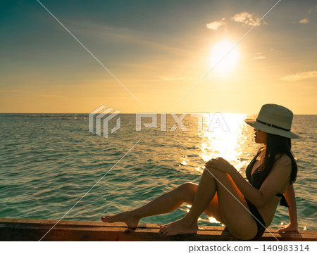 Woman in a bikini relaxing on a wooden pier while watching a golden tropical sunset over calm ocean. Luxury summer vacation, coastal tourism, wellness, seaside leisure, and peaceful travel lifestyle. Woman in a bikini relaxing on a wooden pier while watching a golden tropical sunset over calm ocean. Luxury summer vacation, coastal tourism, wellness, seaside leisure, and peaceful travel lifestyle. 140983314