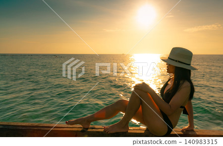 Woman sitting beside sparkling ocean during a breathtaking tropical sunset, overlooking calm sea from wooden pier. Relaxation, luxury travel, summer vacation, coastal wellness, and peaceful lifestyle. 140983315