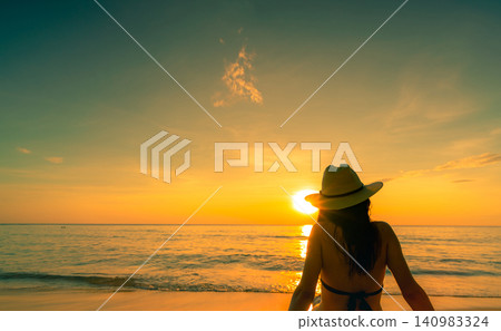 Woman wearing sun hat enjoying tranquil tropical sunset while sitting on beach facing ocean. Luxury vacation, island tourism, golden hour, seaside serenity, relaxation, and inspiring travel lifestyle. Woman wearing sun hat enjoying tranquil tropical sunset while sitting on beach facing ocean. Luxury vacation, island tourism, golden hour, seaside serenity, relaxation, and inspiring travel lifestyle. 140983324
