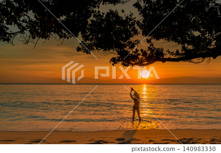 Silhouette of woman stretching in gentle ocean waves beneath a tropical tree at golden sunset. Peaceful beach wellness, summer travel, relaxation, coastal lifestyle, and serene vacation destination. Silhouette of woman stretching in gentle ocean waves beneath a tropical tree at golden sunset. Peaceful beach wellness, summer travel, relaxation, coastal lifestyle, and serene vacation destination. 140983330