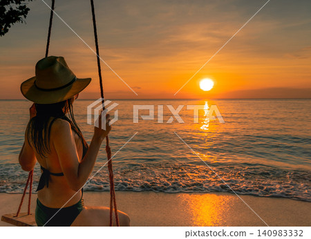 Golden sunset reflecting across the tropical ocean as a woman enjoys beach swing beside gentle waves. Luxury vacation, serene coastal destination, summer travel, relaxation, mindfulness, and wellness. 140983332