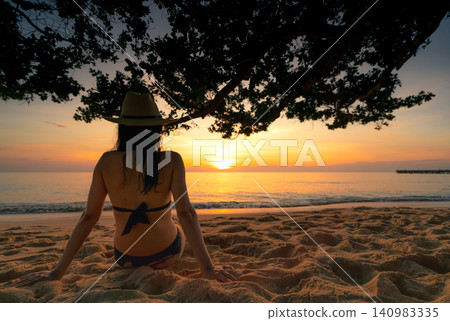 Golden sunset over tropical shoreline with woman relaxing on the sand under a seaside tree. Beach vacation, luxury travel, wellness retreat, summer lifestyle, ocean horizon, and coastal paradise. 140983335