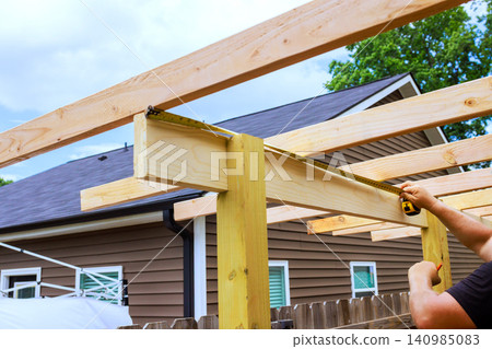 Worker uses tape measure to check length of wooden beam during shed building project behind house. Worker uses tape measure to check length of wooden beam during shed building project behind house. 140985083
