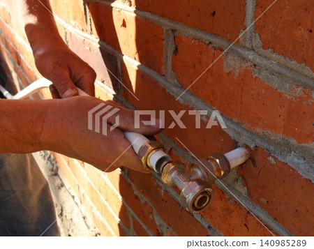A close-up shows the hands of a tradesman installing white metal-plastic pipes using a brass tee fitting. The plumbing installation work is taking place against the backdrop of an exterior red brick w A close-up shows the hands of a tradesman installing white metal-plastic pipes using a brass tee fitting. The plumbing installation work is taking place against the backdrop of an exterior red brick w 140985289