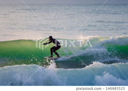 Surfer male riding on glassy wave in ocean with evening sunlight Surfer male riding on glassy wave in ocean with evening sunlight 140985525