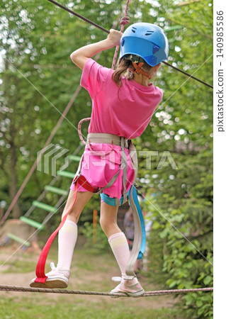 A girl goes through a suspended obstacle course, holding on to a rope. Rope Park of Amusement and Outdoor Activities A girl goes through a suspended obstacle course, holding on to a rope. Rope Park of Amusement and Outdoor Activities 140985586