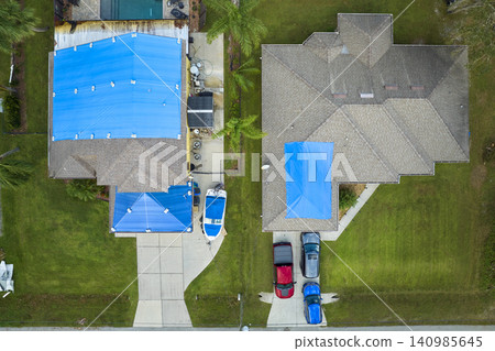 Aerial view of damaged in hurricane Ian house roof covered with blue protective tarp against rain water leaking until replacement of asphalt shingles Aerial view of damaged in hurricane Ian house roof covered with blue protective tarp against rain water leaking until replacement of asphalt shingles 140985645