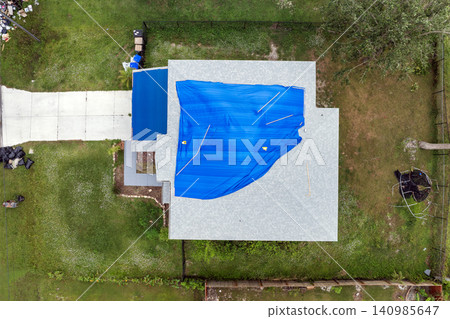 Aerial view of damaged in hurricane house roof covered with blue protective tarp against rain water leaking until replacement of asphalt shingles Aerial view of damaged in hurricane house roof covered with blue protective tarp against rain water leaking until replacement of asphalt shingles 140985647