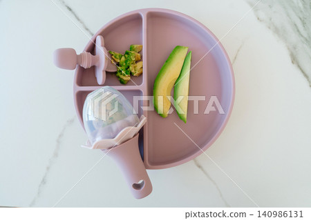 Baby food feeding, in kitchen counter, with baby plate, avocado slices, baby spoon, with plastic texture, from a top-down perspective, with pink, green accents, and nurturing mood. Baby food feeding, in kitchen counter, with baby plate, avocado slices, baby spoon, with plastic texture, from a top-down perspective, with pink, green accents, and nurturing mood. 140986131