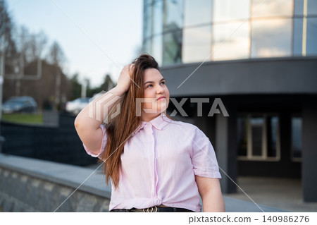 Confident young professional woman in casual attire outside modern glass office building. Corporate lifestyle urban workplace and career success Confident young professional woman in casual attire outside modern glass office building. Corporate lifestyle urban workplace and career success 140986276