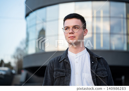 Confident young professional man with glasses standing outside modern glass office building. Business portrait corporate career confidence and workplace setting Confident young professional man with glasses standing outside modern glass office building. Business portrait corporate career confidence and workplace setting 140986283