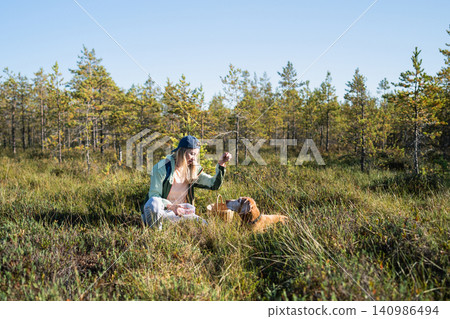 Happy woman hiker collects fresh edible berries sitting at marsh and playing with beloved Vizsla dog Happy woman hiker collects fresh edible berries sitting at marsh and playing with beloved Vizsla dog 140986494