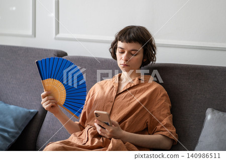 Tired woman using paper fan to cool down in room in summer, using mobile phone. Lack of ventilation Tired woman using paper fan to cool down in room in summer, using mobile phone. Lack of ventilation 140986511
