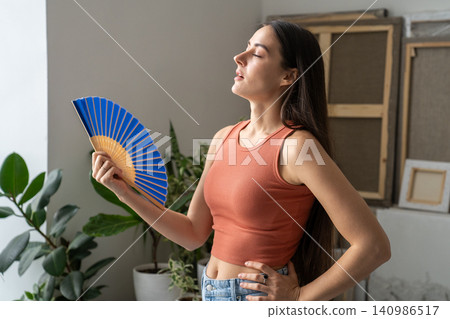 Woman suffering from overheating at home without air conditioner, waving hand paper fan for cooling. Woman suffering from overheating at home without air conditioner, waving hand paper fan for cooling. 140986517