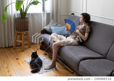 Suffering woman fanning, enduring high temperatures with pet struggling in airless apartment in heat Suffering woman fanning, enduring high temperatures with pet struggling in airless apartment in heat 140986518