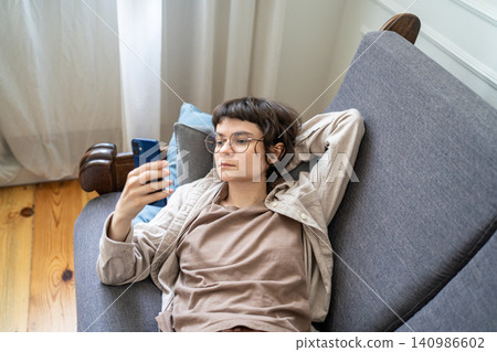 Relaxed woman browsing social media on smartphone while lounging on sofa. Zoomer girl wasted time Relaxed woman browsing social media on smartphone while lounging on sofa. Zoomer girl wasted time 140986602