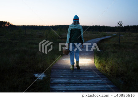 Woman foreger walks through peat bog at wooden path at dusk, lighting her way with headlamp on head Woman foreger walks through peat bog at wooden path at dusk, lighting her way with headlamp on head 140986616