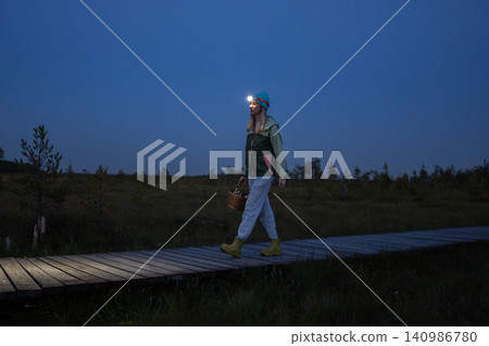 Woman foreger walks through peat bog at wooden path at dusk, lighting her way with headlamp on head Woman foreger walks through peat bog at wooden path at dusk, lighting her way with headlamp on head 140986780