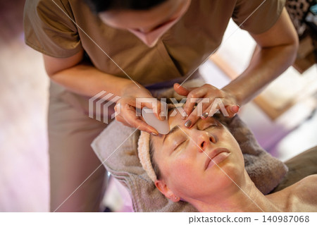Cosmetologist in salon gently scrubbing woman client face skin using Gua sha massage stone scrubber. 140987068