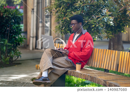 Smart confident african american man freelancer working online on laptop sitting on park bench. Smart confident african american man freelancer working online on laptop sitting on park bench. 140987080