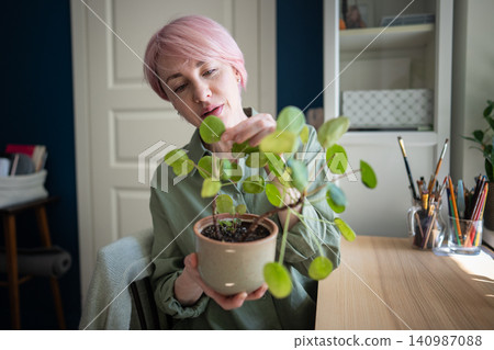 Mature woman home gardener checking potted Pilea peperomioides plant foliage. Green hobby. Mature woman home gardener checking potted Pilea peperomioides plant foliage. Green hobby. 140987088