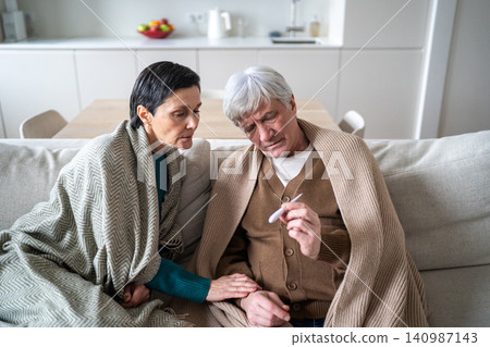 Old covered in blankets checking temperature level with thermometer sitting on sofa at home Old covered in blankets checking temperature level with thermometer sitting on sofa at home 140987143