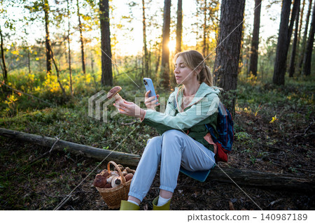 Woman hiker resting, sitting on log in forest, taking pictures of big boletus in hand on smartphone. Woman hiker resting, sitting on log in forest, taking pictures of big boletus in hand on smartphone. 140987189