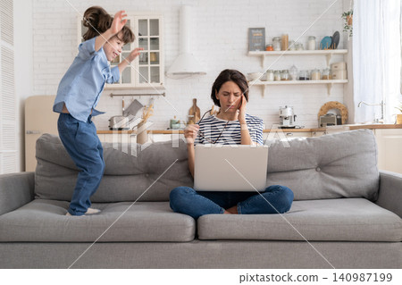 Tired mother sit on couch, work on laptop at home, hyperactive little kid jumping drawing attention. Tired mother sit on couch, work on laptop at home, hyperactive little kid jumping drawing attention. 140987199