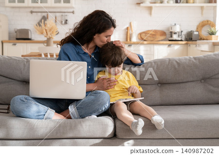 Remote worker mom or freelancer embrace kid with tablet using laptop computer to work from home Remote worker mom or freelancer embrace kid with tablet using laptop computer to work from home 140987202