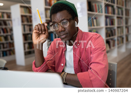 Focused African American student guy in glasses studying sitting in library watching online lesson Focused African American student guy in glasses studying sitting in library watching online lesson 140987204