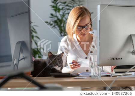 Woman using calculator, planning expenses while working on desktop from home, cat nearby on table Woman using calculator, planning expenses while working on desktop from home, cat nearby on table 140987206