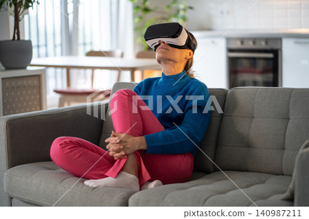 Intrigued European woman wearing virtual reality glasses relaxing after day at work sits on sofa 140987211