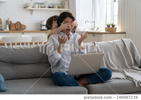 Mom work on laptop at home during lockdown, child distracts from work, covering her mother's eyes Mom work on laptop at home during lockdown, child distracts from work, covering her mother's eyes 140987239