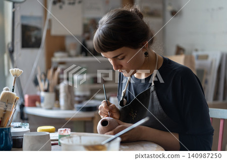 Lady master paints clay jug with serious and concentrated expression sitting at table in workshop Lady master paints clay jug with serious and concentrated expression sitting at table in workshop 140987250