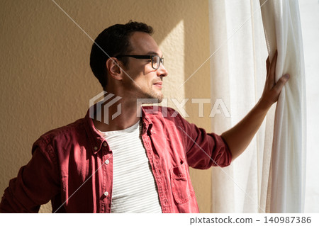 Meeting new day. Young pensive man opening curtains, looking through window enjoying sunlight Meeting new day. Young pensive man opening curtains, looking through window enjoying sunlight 140987386