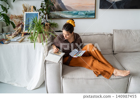Professional craftswoman drawing picture in sketchbook with watercolor lying on couch in living room Professional craftswoman drawing picture in sketchbook with watercolor lying on couch in living room 140987455