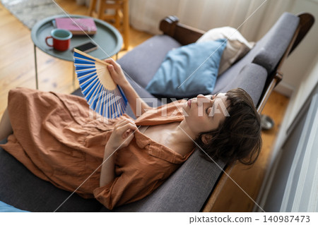 Stressed woman suffering from heat at home sitting on sofa waving paper fan with closed eyes. Stressed woman suffering from heat at home sitting on sofa waving paper fan with closed eyes. 140987473