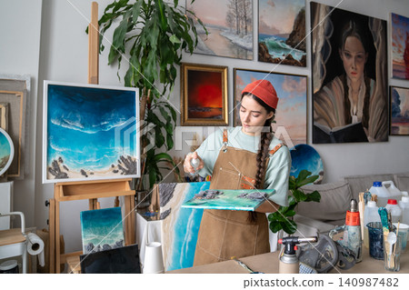 Concentrated craftswoman works on epoxy painting, spraying cleaning liquid on canvas with resin art. Concentrated craftswoman works on epoxy painting, spraying cleaning liquid on canvas with resin art. 140987482