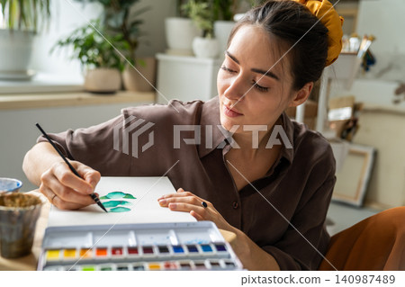 Pensive woman artist enjoying new watercolor paints, drawing sketches in sketchbook at home studio. 140987489