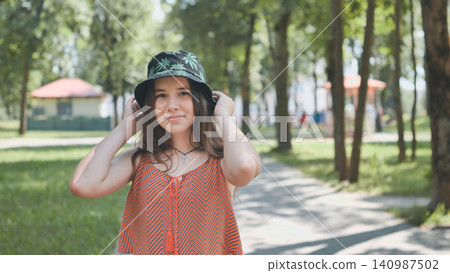 Young woman wearing trendy bucket hat, walking through sunlit park with lush greenery, basking in warm summer afternoon 140987502