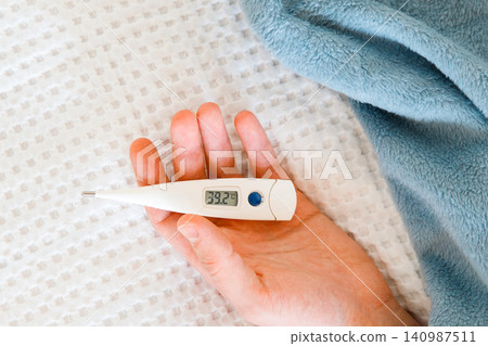 Closeup of man hand holding digital thermometer, lying on bed. High body temperature, flu Closeup of man hand holding digital thermometer, lying on bed. High body temperature, flu 140987511
