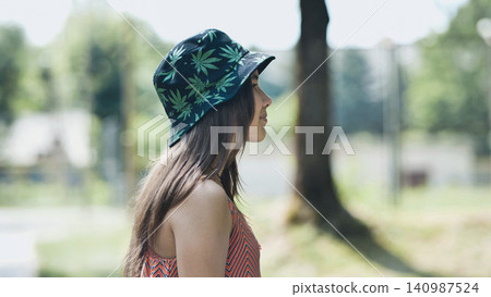 Young female relaxing in green park, wearing trendy bucket hat with marijuana leaf pattern, enjoying sunny afternoon 140987524
